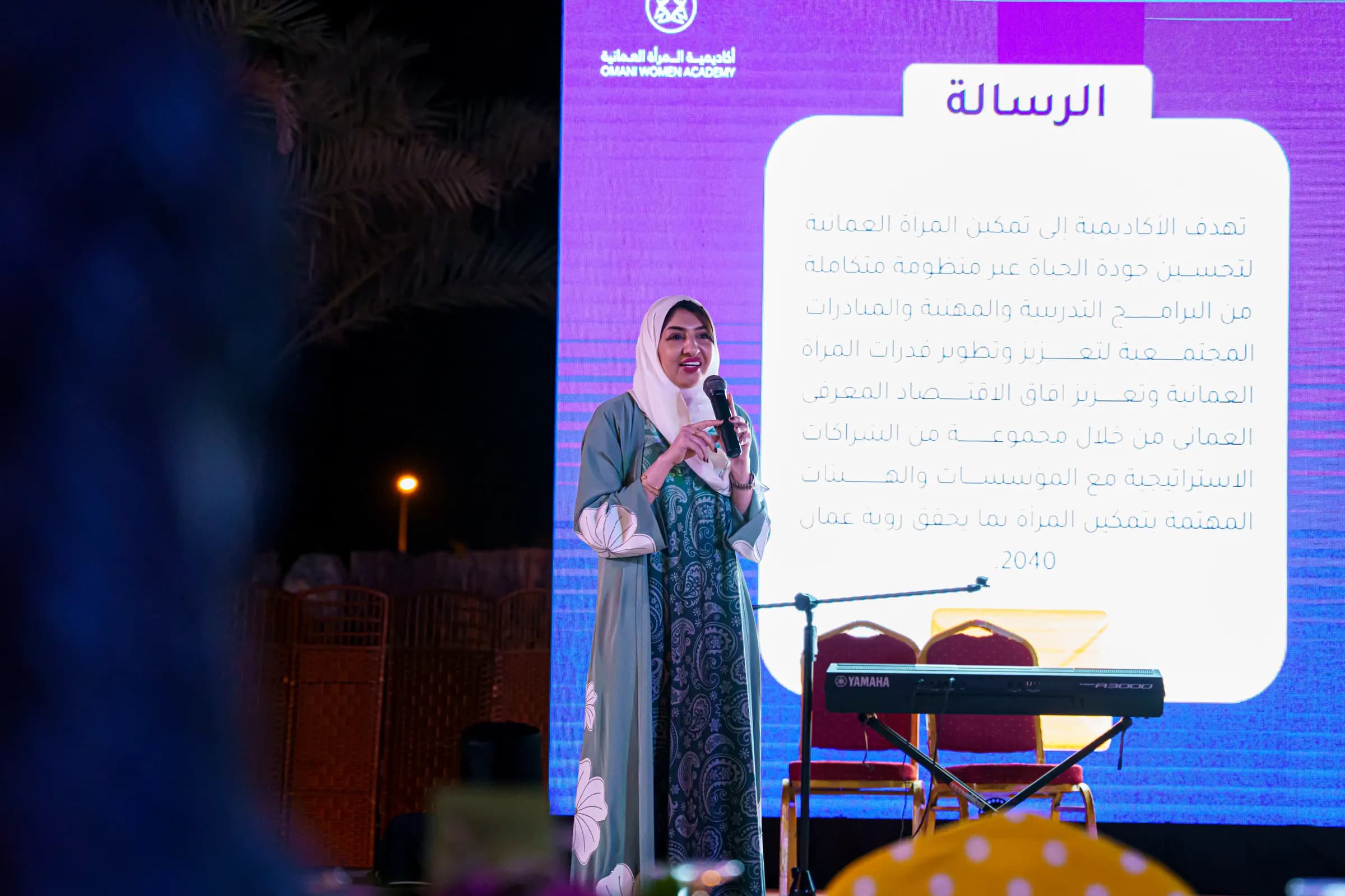 متحدثة على المنصة تحمل الميكروفون أمام شاشة تعرض رسالة الأكاديمية